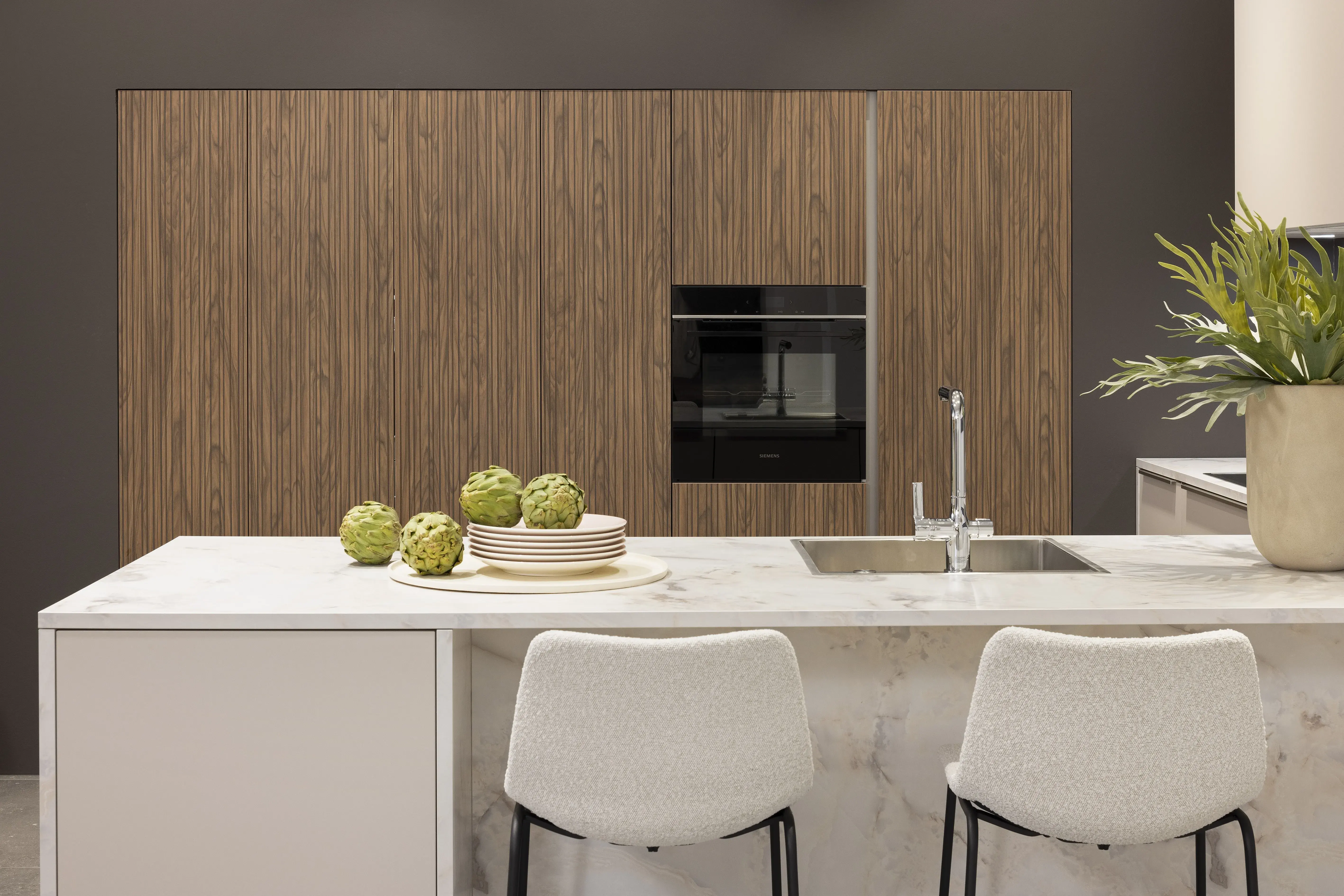 APL: Marmor Bianco 25 mm NB Front: Feel Sahara softmatt Front: WArtline Stripe Wood Choco Korpus: Sahara und Graphit Griff: Kehlleiste Edelstahloptik, und Griffe Edelstahloptik Geräte: Siemens  Armatur: Reginox Spüle: Reginox   Features: eingekoffertes Schrankelement, Tür zum HWR in Frontmaterial, niederer Sockel, Hängeoberschränke asymmetrisch mit Dunstabzug (als Esse geplant), Ecklösung mit LeMans Auszug, Theke mit Essplatz, Ordnungssystem Holz in Auszügen, Downlight in Arbeitsplatzbeleuchtung 